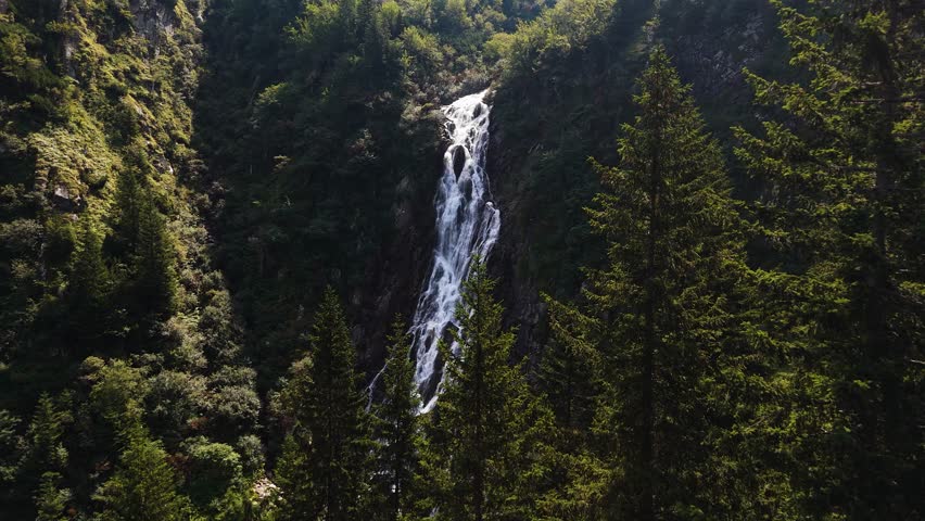 Drone flies steadily above treetops toward waterfall in mountain forest
