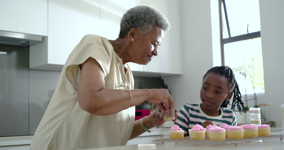 Grandma is picking sprinkles and teaching grandkid as they are decorating pink cupcakes in kitchen. Family, baking, teamwork, mentoring, multigenerational, dessert, colorful
