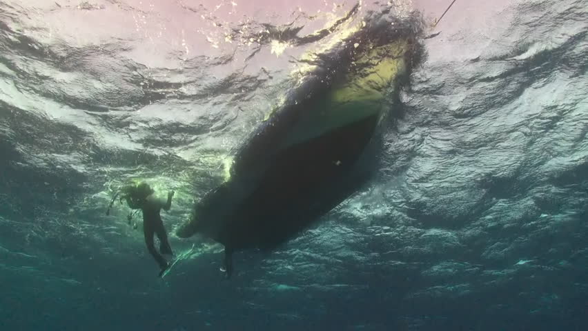 A diver descends into the clear blue waters of La Palma, Canary Islands. Sunlight filters through the surface as they explore the underside of a boat, a hidden world beneath the waves.