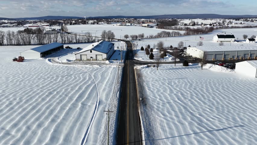 Aerial view of rural American landscape in winter with snow covered fields, farm buildings and road cutting through countryside. Calm seasonal scene under clear daylight. Pennsylvania, USA.