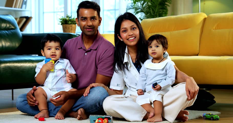 Indian Asian twin infant babies smiling and looking at camera while sitting with young parents on carpet surrounded by toys in modern home living room, capturing joyful family portrait and bonding