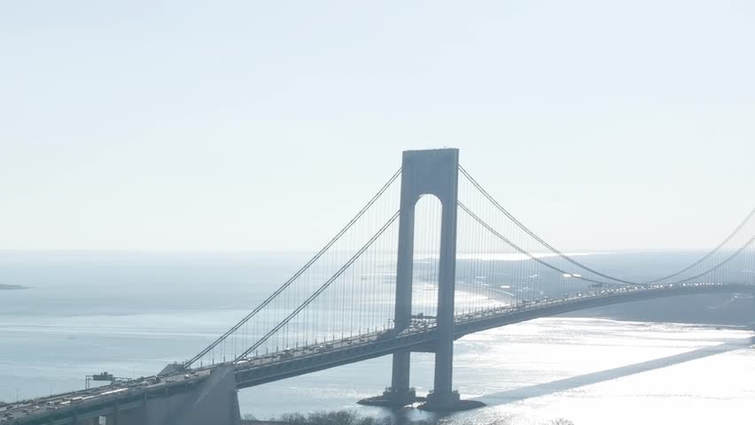 Traffic crosses the Verrazzano Narrows Bridge in New York. The bridge connects Staten Island and Brooklyn. The sun reflects off the water on a clear day.