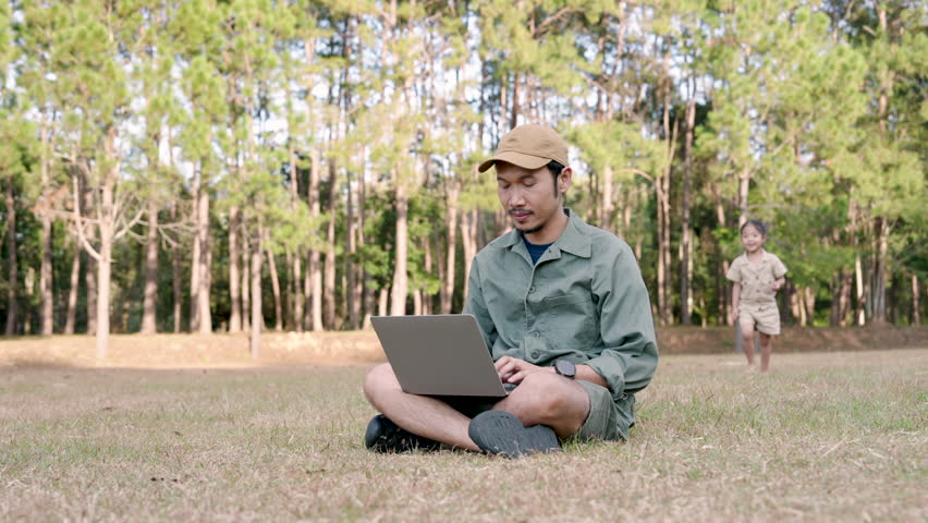 Asian adult father smiling while daughter hugs from behind sitting on dry grass using laptop in national park showing joyful family connection during relaxing vacation in outdoor nature environment