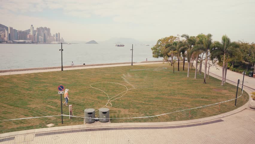 Wide static shot of a worker lifting a sprinkler from the ground and spraying water across a grass lawn in a waterfront park with Hong Kong skyline and harbor in background under daylight