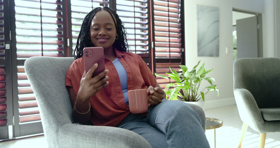 Adult African woman reacting to message, holding pink phone and mug, scrolling and laughing at home. Armchair, device, cup, braided, shutters, plant, lounge