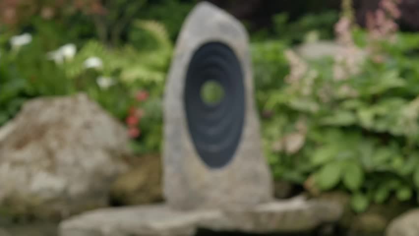 A large stone sculpture with concentric oval openings stands on a rock in a peaceful garden. Green plants and flowering blooms surround the stone art near a water feature, encouraging quiet reflection