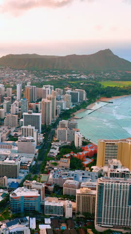 High angle view of urban buildings and the coastline with a mountain silhouette under a soft evening sky. Sunset aerial view over Honolulu city and Diamond Head. Vertical video.