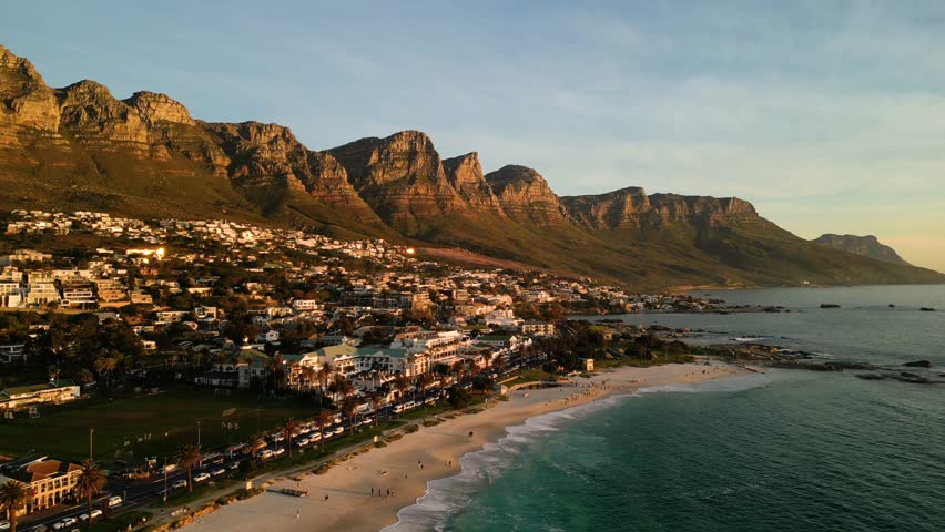 Aerial drone footage flying away from Camps Bay in Cape Town South Africa during golden hour showing coastal landscape and warm sunset light.