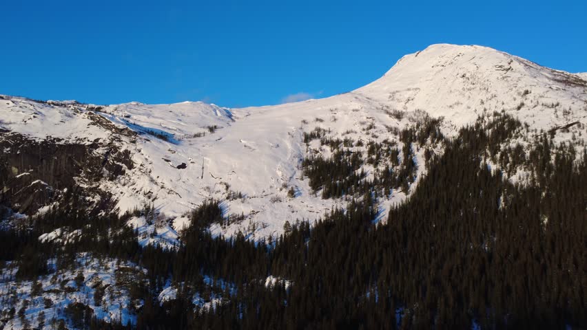 Drone flying away from snow covered mountain and forest in Norway. Aerial view of winter alpine landscape and Scandinavian wilderness.