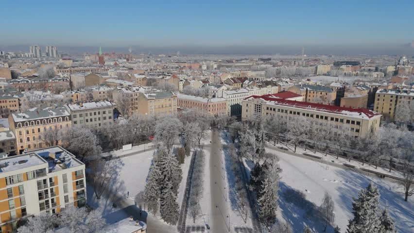 Drone Aerial View Of Riga City Skyline On A Sunny Cold Winter Day With Snow Covered Rooftops, Smoking Chimneys, Clear Blue Sky And The Scenic Ziedondarzs Park In The Heart Of The Latvian Capital