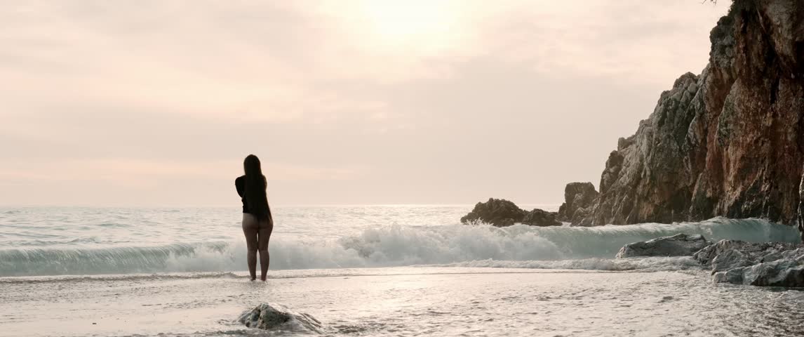 Cinematic anamorphic wide shot of a woman standing barefoot in shallow sea water at sunset. Waves wash around her legs as she faces the horizon near dramatic rocky cliffs, then slowly walks out of frame.