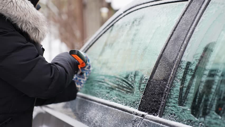 Person in winter gloves scrapes frost from a vehicle side window with an ice scraper, clearing the glass for visibility and safe driving in cold weather.