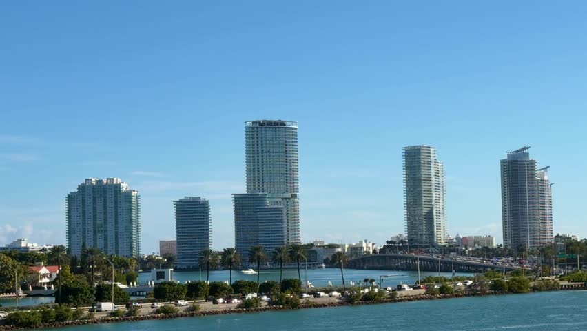Miami South Beach skyline featuring modern buildings, numerous tropical palm trees, cars driving on E Port Boulevard, and boats docked in the blue water on a sunny day
