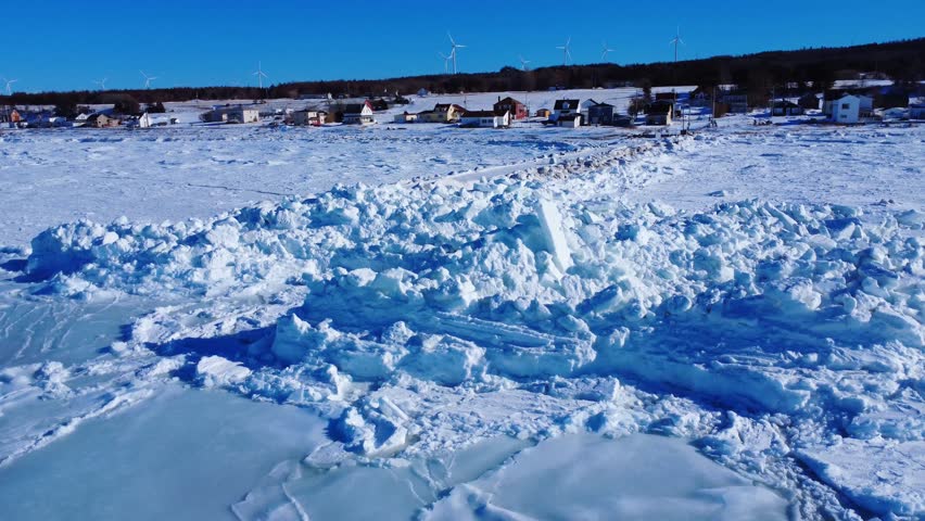 The shoreline of the coastal village of Baie-des-Sables is buried under a mass of ice chunks piled up by the tides of the St. Lawrence River, with wind turbines in the background under a blue sky. Quebec, Canada, Winter 2026.