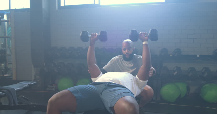 African male trainer spotting trainee at gym bench with dumbbells lowering, pressing for power. Weightlifting, backlit, industrial, coaching, muscular, athletic, focus