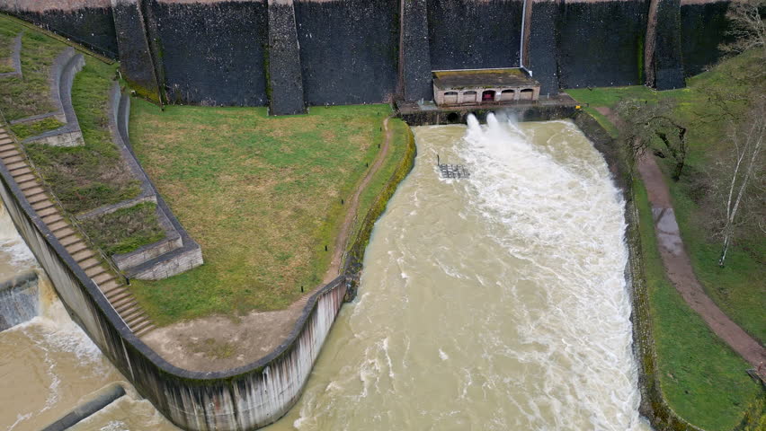 drone view of dam spillway and outlet pipes releasing water into river below, with reservoir lake behind and forested shoreline surrounding it