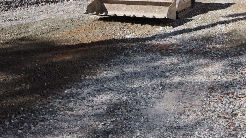 Construction vehicle uses blade to smooth gravel on road surface at work site.