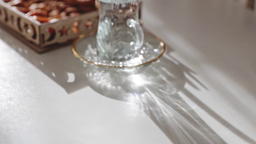 A glass of clean water sits on a white table in a saucer, along with a carved wooden tray of dried fruit. Close-up, side view, slow motion, and forward tracking. Concept: festive food and drinks, Ramadan Kareem, Eid Mubarak.