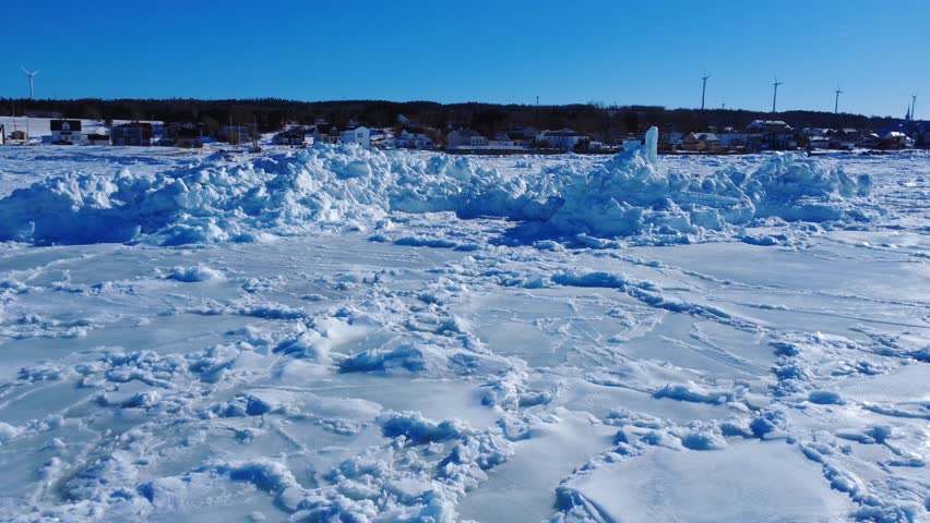 A wall of ice, compacted by the tides, rises in front of the houses of a coastal village, with wind turbines against a blue sky in the background. Baie-des-Sables, Gaspé Peninsula, Quebec, Canada, 2026.