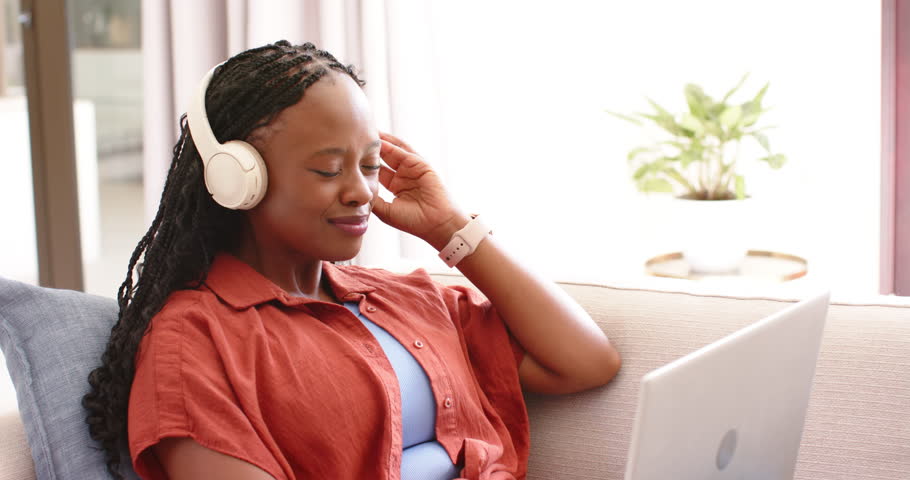 Adult African woman wearing rust top sitting on sofa touching white headphones, laughing at laptop. Cozy, daylight, home, plant, window, curtains, relaxed