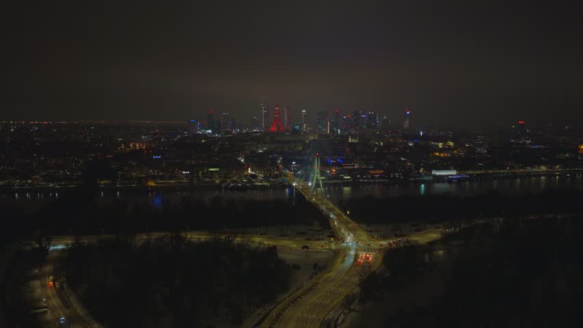 Aerial night scene of Warsaw shows Swietokrzyski Bridge, Vistula River, and the red lit Palace of Culture and Science, with traffic light trails and neon reflections on water.