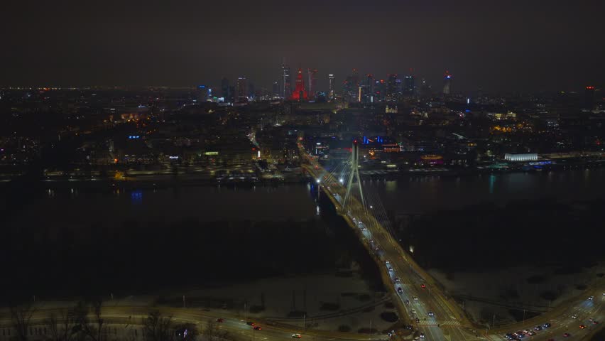 Aerial night view approaches Swietokrzyski Bridge in Warsaw as traffic moves, city skyline glows with Palace of Culture and Science, lights reflect on river, winter snow shows.