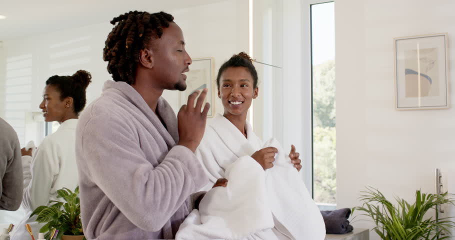 African American couple in robes post-wash standing by bathroom sink drying faces with white towels. Home, sunlit, modern, duo, grooming, selfcare, intimate