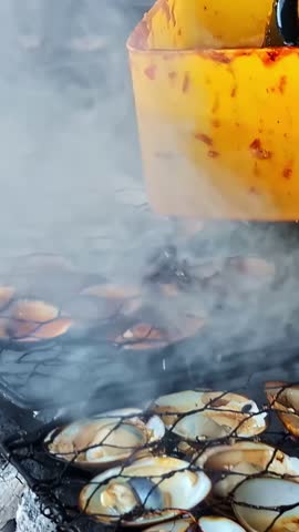 Fresh clams grilling over an open charcoal fire with smoke rising through a metal grate