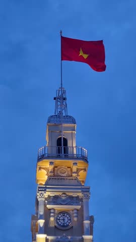 Vertical shot of the Vietnamese flag flying atop the City People’s Committee Building tower in Ho Chi Minh City during early night hours, under an overcast sky and calm urban atmosphere