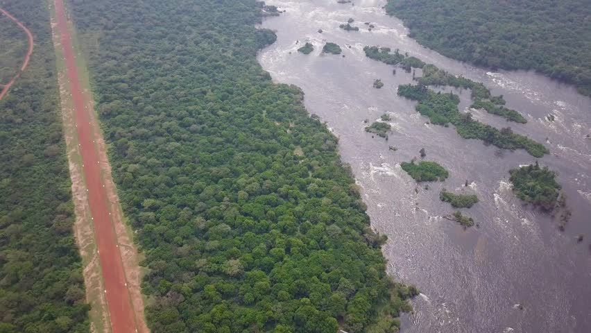 Remote dirt airstrip running along the Nile River through dense savanna woodland in Murchison Falls National Park, Uganda, showing wilderness access and safari logistics, aerial wide shot.