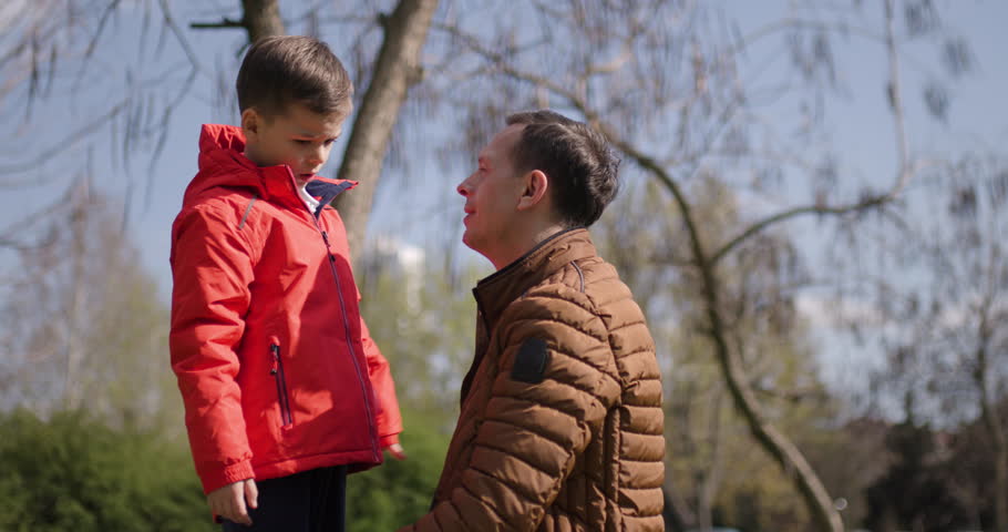A joyful moment unfolds as a father embraces his son in a park filled with trees. The scene captures the warmth of family love under a clear blue sky.