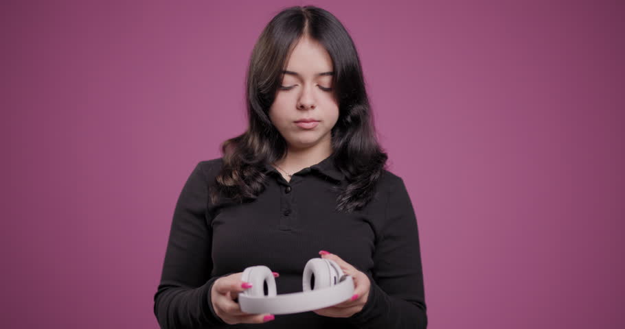 A focused young woman in a black shirt carefully examines a pair of white wireless headphones, set against a soft pink background.