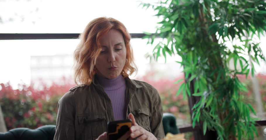 A woman with auburn hair is sitting at a table in a cafe during the day. She is interacting with her mobile device, with a leafy plant nearby and a bright window in the background.