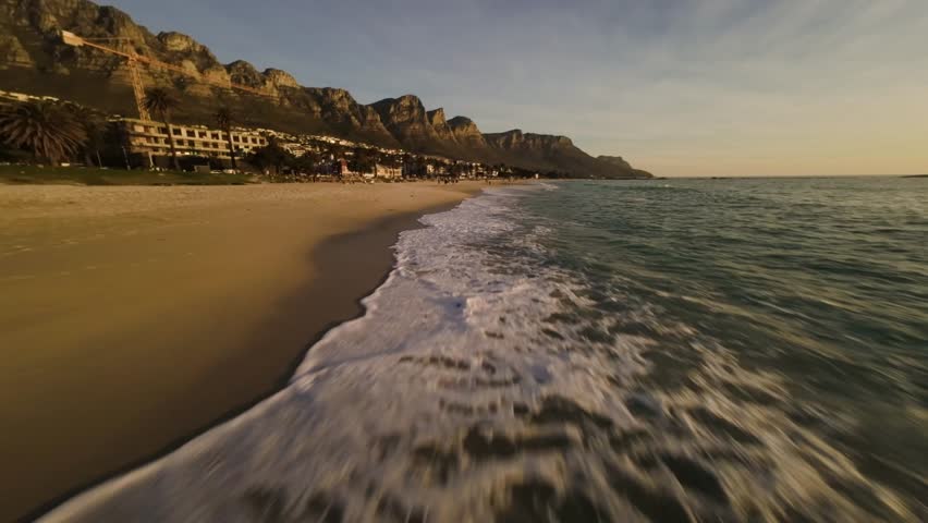 Smooth FPV drone footage flying straight along Camps Bay beach in Cape Town South Africa capturing ocean waves and golden sunset light.