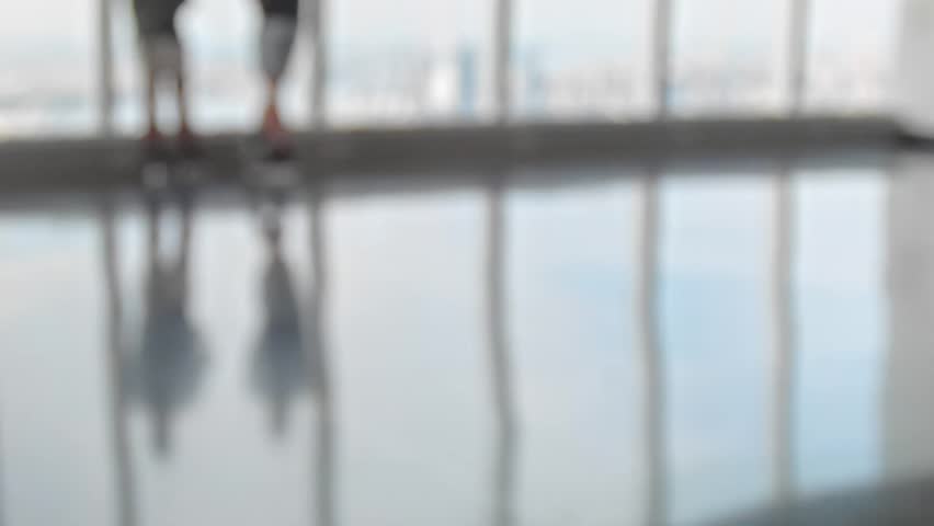 Unknown defocused people walk around inside a brightly lit room with reflective floor and large windows, persons wear casual clothes, shorts and summer outfits.