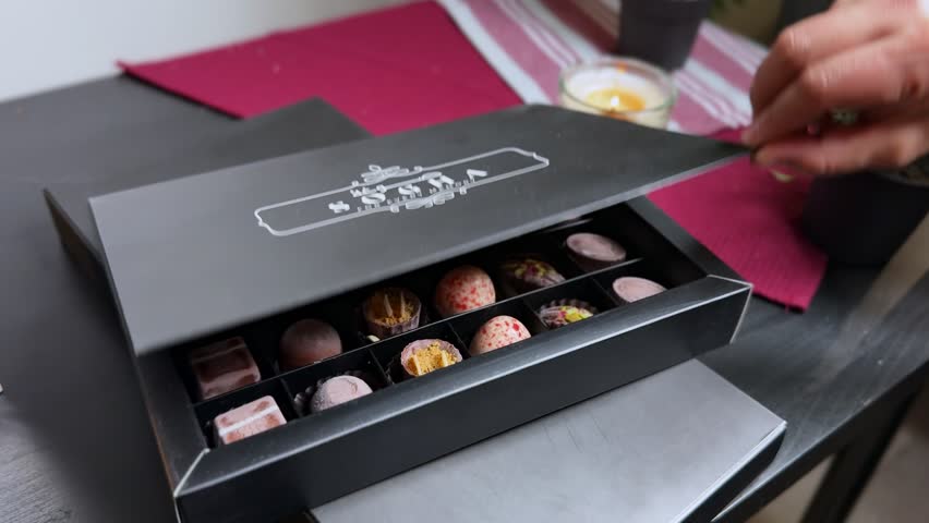 Close-up of assorted gourmet chocolates in an open black gift box on an indoor table, with a lit candle and red runner creating a cozy, festive dessert mood.