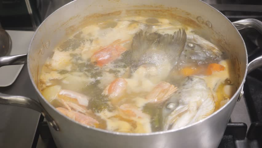 Seafood broth simmers in a pot with fish head, shrimp shell (Penaeus spp.), onion (Allium cepa), and herbs such as bay leaf (Laurus nobilis), creating a rich stock base, close up shot.