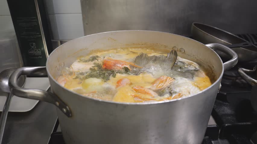 Close view of a pot of seafood stock simmering on the stove, with a fish head, shrimp shells, onion pieces, and herbs visible in bubbling broth, preparing a flavorful base for soup or sauce.