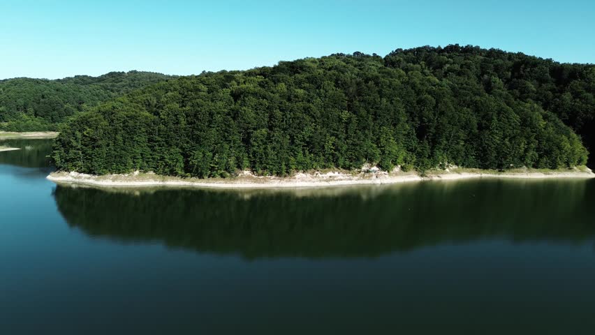 Aerial view of calm Garasi Lake surrounded by dense green forest under clear blue sky during summer in Serbia, captured from a drone above still water and shoreline.