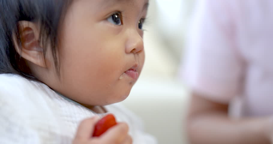 Asian toddler girl holding strawberry piece with tiny fingers, looks down softly, crumbs on hand, warm natural light, close up moment, peaceful scene, child concentration, early development learning