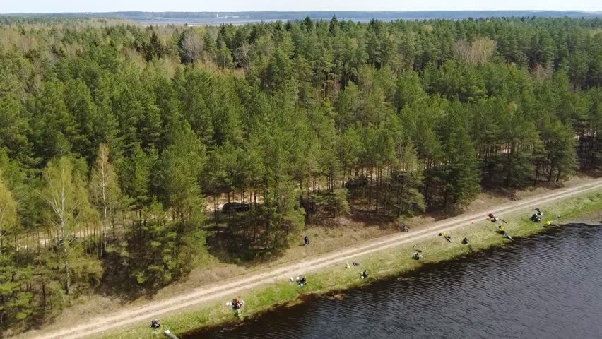 Groups of people are seen fishing along the riverbank while surrounded by tall trees in a forest area. The sun is setting, and the water reflects the light.