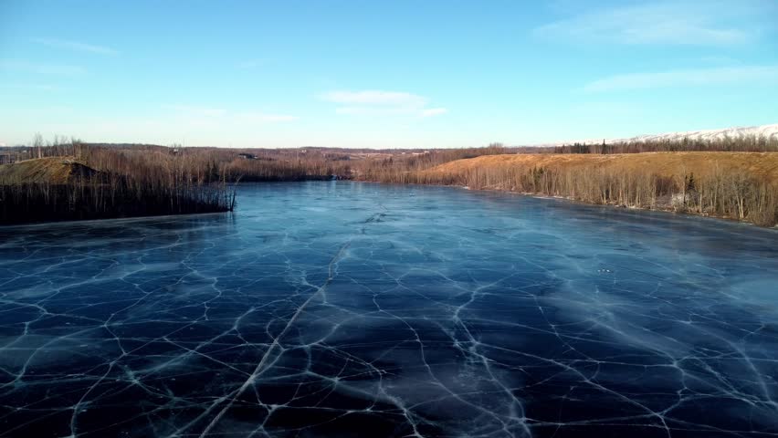 Low level drone flight skimming twenty to thirty feet above the frozen surface of Matanuska Lake, traveling in the opposite direction across the vast expanse. Deep blue ice with striking gray highligh