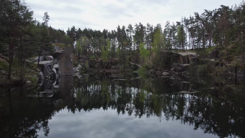 Small scenic lake with steep rocky shores among the pine forest in springtime, aerial view while moving forward
