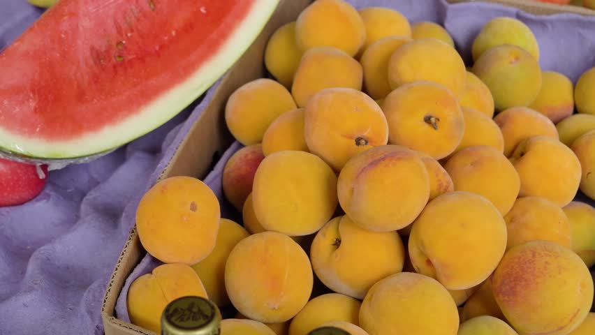 Close-up of apricots stacked in a box at a market stall with other produce nearby.