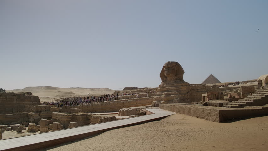 Wide angle handheld real-time footage of the Great Sphinx with the Pyramids of Giza visible in background under clear blue sky. Tourists visit ancient Egyptian landmark on the Giza Plateau