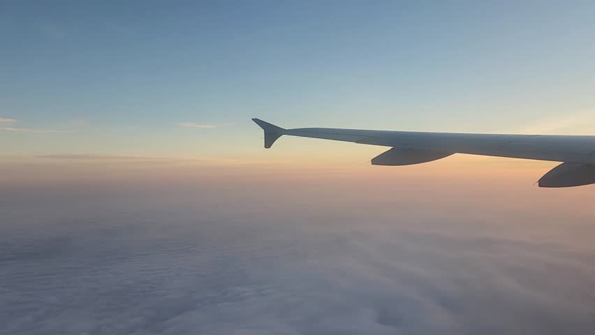 Golden sunset light floods the sky as layered clouds drift beneath the plane window, painting the horizon in warm orange and soft violet hues above the endless sky