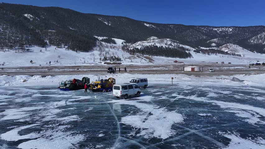 A cinematic pull-out drone shot reveals hovercrafts and vehicles on the vast, crystalline frozen Lake Baikal under a clear blue sky, highlighting the majestic winter landscape.