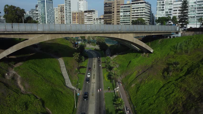 Aerial shot of a bridge over a road called "Bajada Balta". Done flies backwards and up shoting the coastal road named "Costa Verde" and the many buildings of the Miraflores district of Lima, Peru.