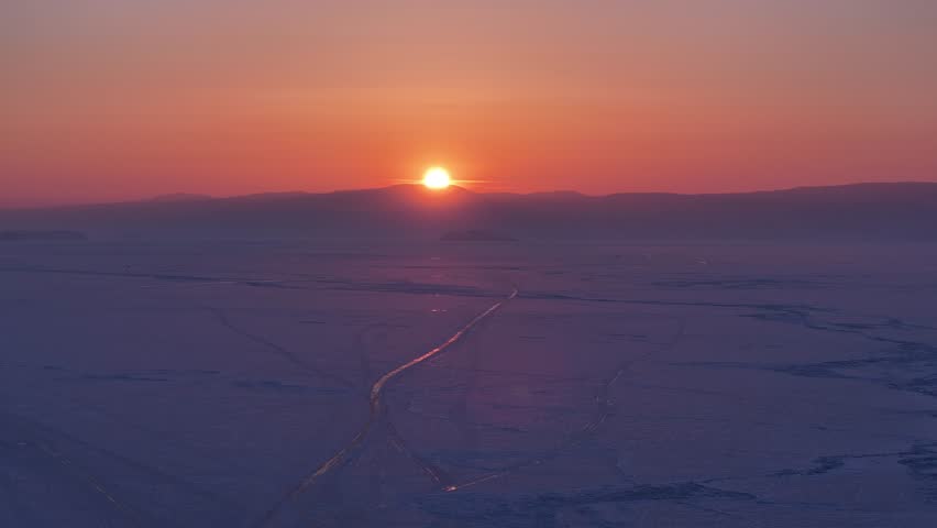 Serene aerial push-in shot over the vast frozen surface of Lake Baikal at sunset, showcasing intricate ice cracks and distant mountains.