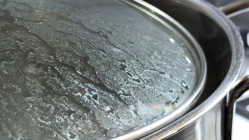 Water boils intensely inside a stainless steel pot covered with a glass lid. The trapped steam condenses into droplets as the water gurgles and splashes against the surface.